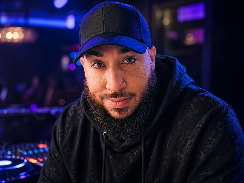 DJ Angel in a black cap and hoodie in front of a lit DJ booth