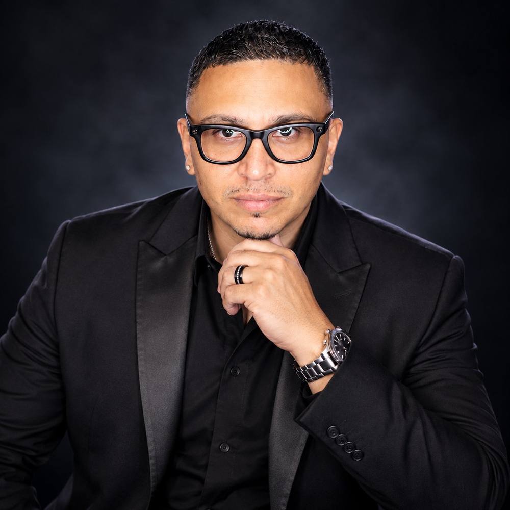 Portrait of DJ Mario in a black suit and glasses against a dark backdrop